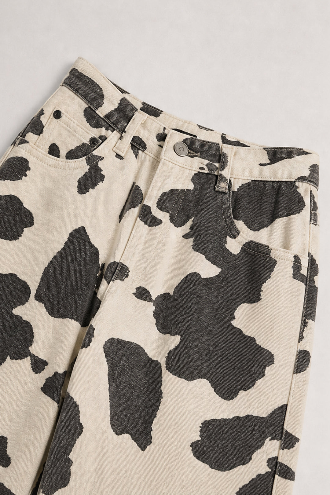 Close-up of a pair of pants with a black and off-white cow print pattern, showing the waistband, belt loops, and front pockets on a light gray background.