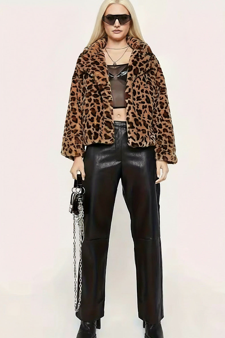 A blonde model wearing a brown and black leopard print faux fur jacket, black leather pants, a sheer black crop top, and black boots, holding a black handbag with a silver chain strap against a light beige background.
