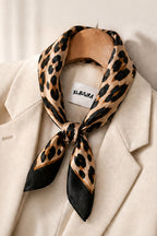A leopard print silk scarf with black satin ends, tied around a beige blazer on a wooden hanger. The brand label 'KLAUWRA' is visible inside the collar.
