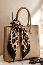 A structured beige tote bag with dual top handles, decorated with a large leopard-print silk scarf tied in a bow at the front. The scarf has black leopard spots on a tan background with solid black lower corners.