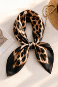 A leopard print silk scarf tied in a bow, displayed on a light beige surface with a straw hat and gold chain partially visible in the background.