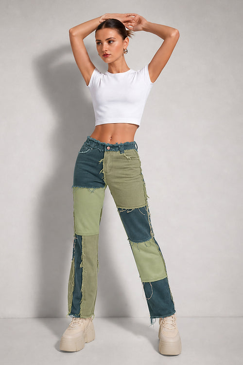 A model wearing a white crop top and a pair of high-waisted, patchwork-style distressed jeans in shades of teal and sage green, paired with beige platform sneakers, against a light gray studio background.