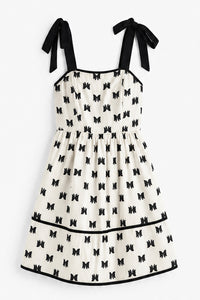A sleeveless white dress with a black bow pattern, black shoulder ties, and black trim, shown flat against a light gray background.