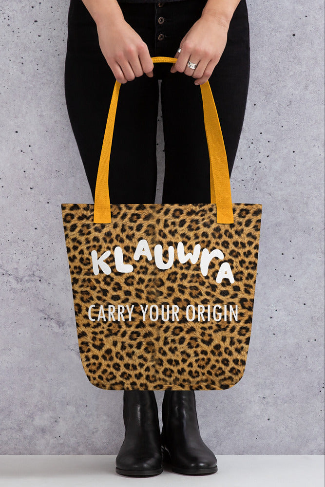 A person wearing black pants and black boots holds a leopard-print tote bag with yellow handles. The bag has white text that reads 'KLAUWRA' and 'CARRY YOUR ORIGIN'.