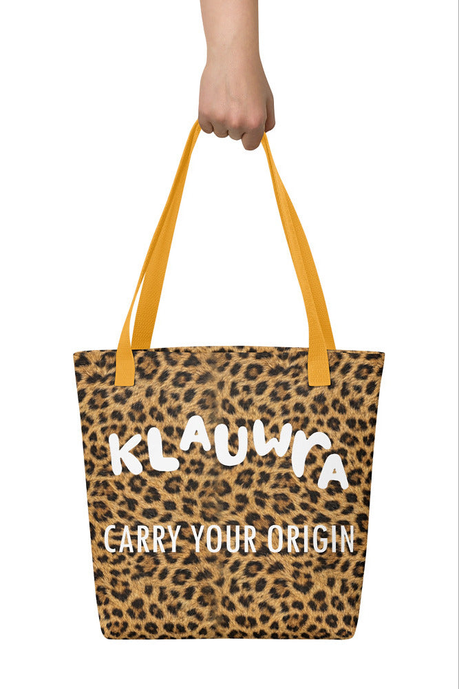 A leopard print tote bag with mustard yellow handles, held by a hand against a white background. The bag features the text 'KLAUWRA' and 'CARRY YOUR ORIGIN' in white.