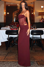 A woman modeling a floor-length burgundy evening gown with a sheer lace bodice and side cutouts, standing in a dimly lit restaurant or event space with tables and chairs in the background.