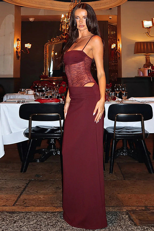 A woman modeling a floor-length burgundy evening gown with a sheer lace bodice and side cutouts, standing in a dimly lit restaurant or event space with tables and chairs in the background.