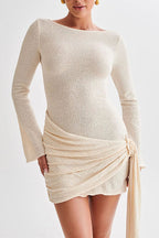 A woman wearing a textured cream knit long-sleeve mini dress with a draped wrap-style skirt and tie detail, shown against a plain light background.