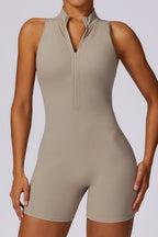 A sleeveless, high-neck athletic romper in a light taupe-beige color, featuring a half-zip neckline, modeled on a person against a light gray studio background.