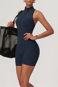A woman modeling a navy blue sleeveless biker-style jumpsuit with a mock neck and front zipper, holding a black mesh tote bag against a light gray studio background.
