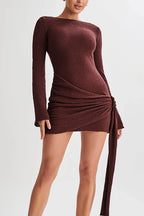 A woman modeling a long-sleeve, off-the-shoulder mini dress in a textured, metallic deep burgundy-brown fabric. The dress features a ruched wrap-style skirt with a side tie, photographed against a clean, light gray studio background.