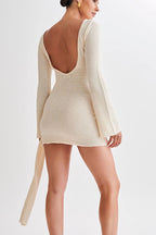A woman modeling a cream-colored, long-sleeve mini dress with a deep open back and a matching tie detail at the waist, shown from a rear three-quarter angle against a plain light background.