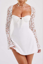 A woman wearing a white mini slip dress with long lace sleeves and a sweetheart neckline with a front tie, photographed against a light gray studio background.