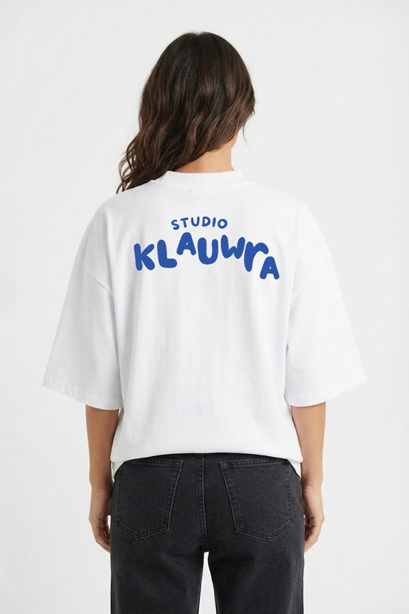 A woman viewed from the back wearing an oversized white T-shirt with 'STUDIO KLAUWRA' printed in blue across the back, paired with dark gray jeans, against a plain white studio background.