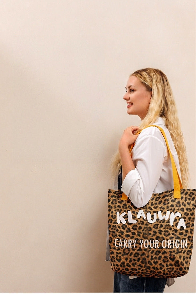 A woman in a white shirt and jeans stands in profile holding a leopard-print tote bag with yellow straps. The bag has the text 'KLAUWRA' and 'CARRY YOUR ORIGIN' printed on it.