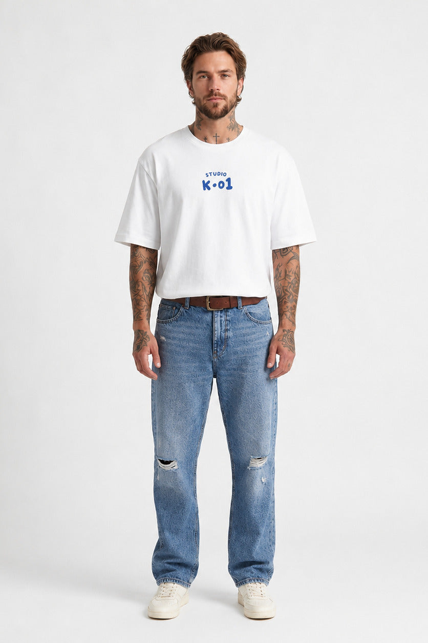 A man modeling a white short-sleeve t-shirt with a blue graphic print on the chest, paired with distressed blue jeans and white sneakers, against a plain light gray background.