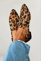 A pair of pointed-toe flat loafers with a leopard print pattern and silver buckle straps, shown on a person's feet wearing blue jeans against a plain light gray background.
