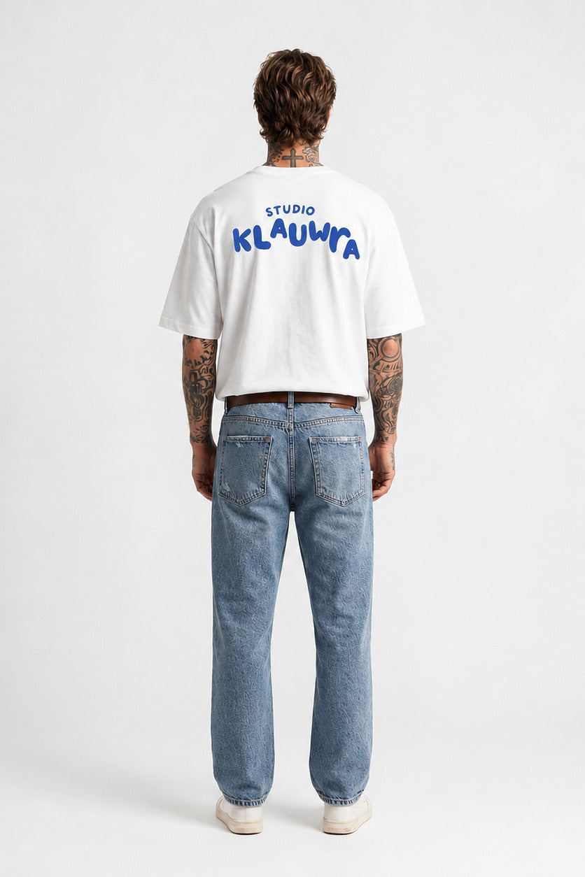 A man viewed from behind wearing a white cropped t-shirt with blue 'STUDIO KLAUWRA' text on the back, paired with light blue jeans and white sneakers, against a plain white studio background.