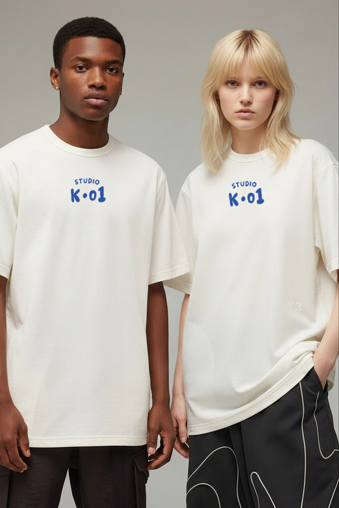 A man and a woman modeling matching off-white oversized T-shirts with blue 'STUDIO K-01' text on the chest, photographed against a plain light gray studio background.