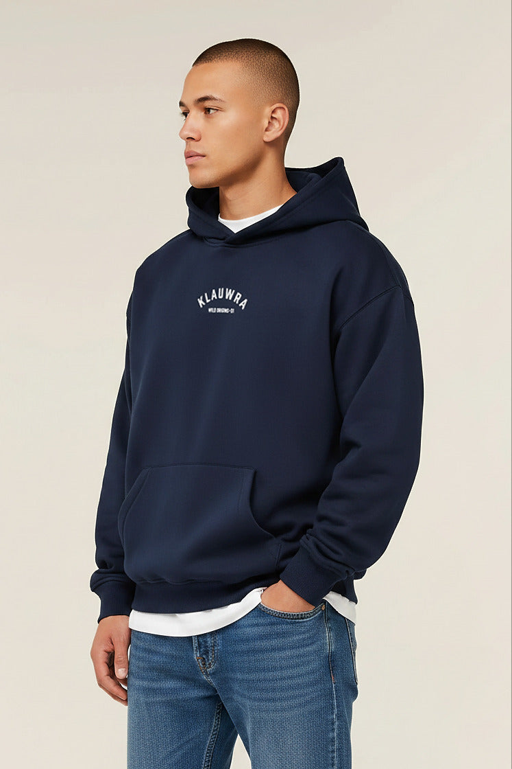 A man wearing a navy blue pullover hoodie with a white arched 'KLAUWRA' logo on the chest, layered over a white t-shirt and blue jeans, photographed against a solid light beige background.
