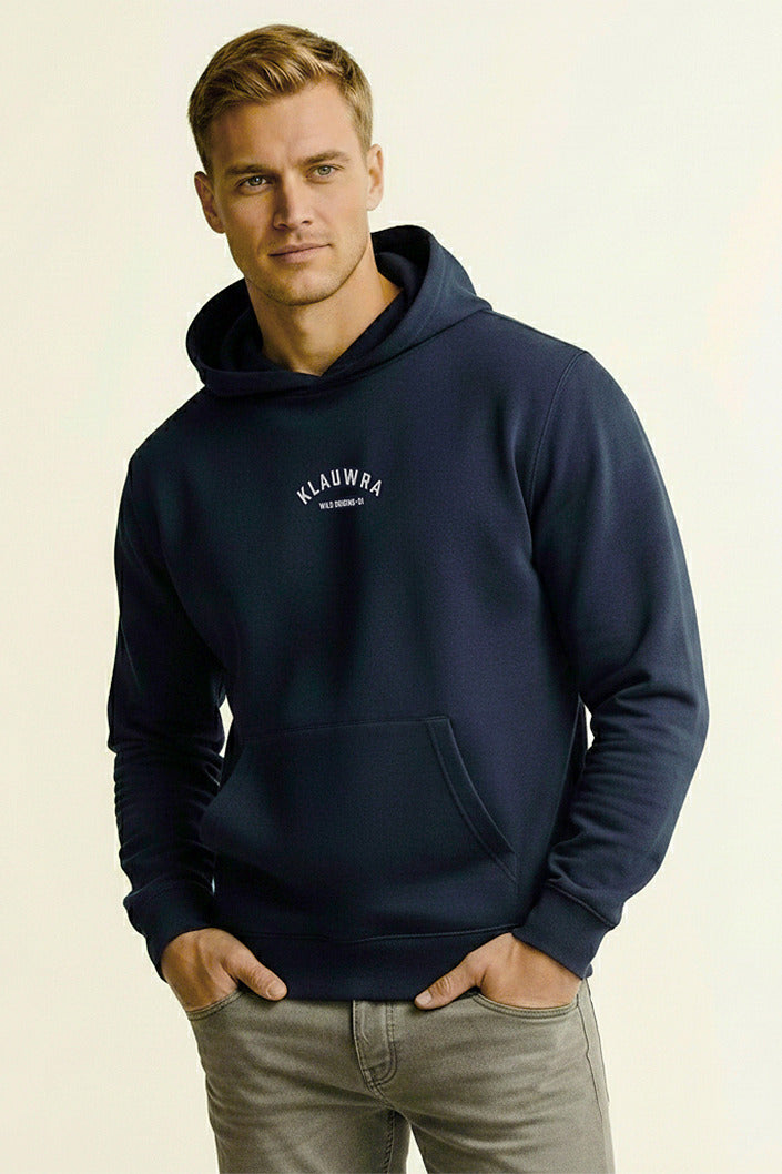 A man wearing a navy blue pullover hoodie with a white 'KLAUWRA' logo on the left chest, paired with gray jeans, against a plain light beige background.