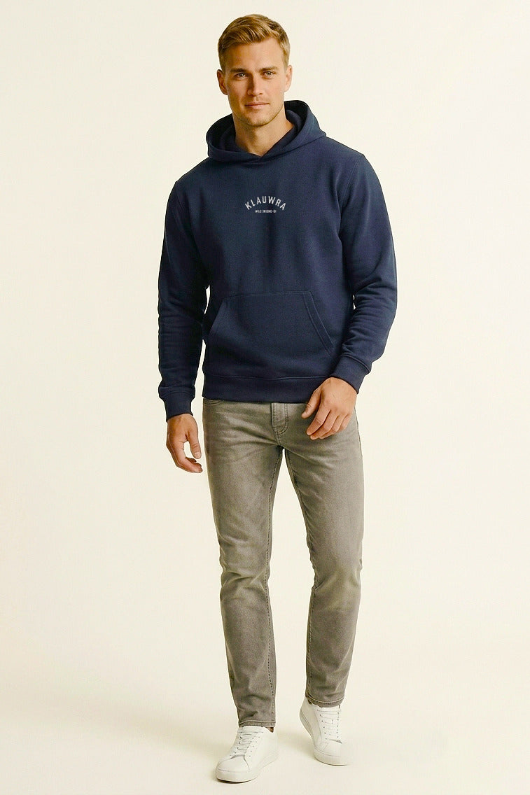 A man modeling a navy blue hoodie with a white 'KLAUWRA' logo on the chest, paired with gray jeans and white sneakers, against a plain off-white studio background.