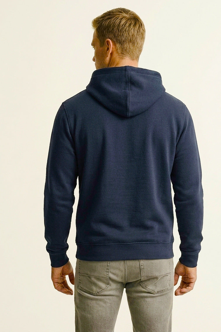 A man viewed from the back wearing a navy blue pullover hoodie and light gray jeans, standing against a plain off-white studio background.