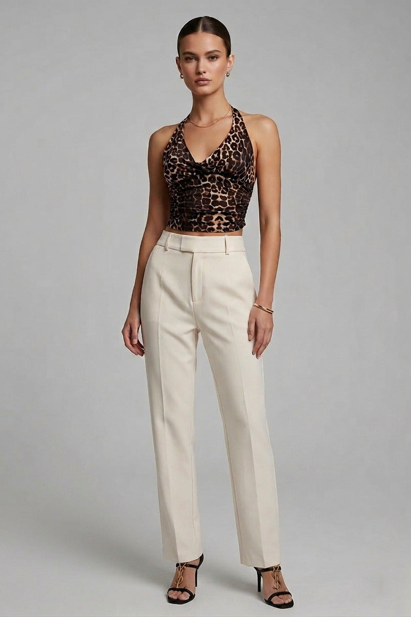 A model stands against a light gray studio background wearing a sleeveless leopard-print halter crop top and high-waisted cream wide-leg trousers with black strappy heels.